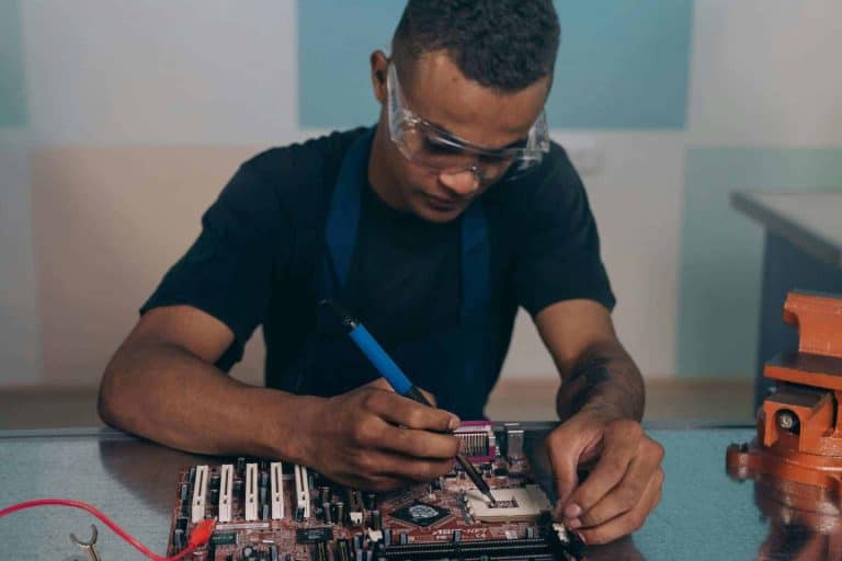 A technician wearing safety glasses works intently on a computer motherboard with a soldering iron. AI Data Centers