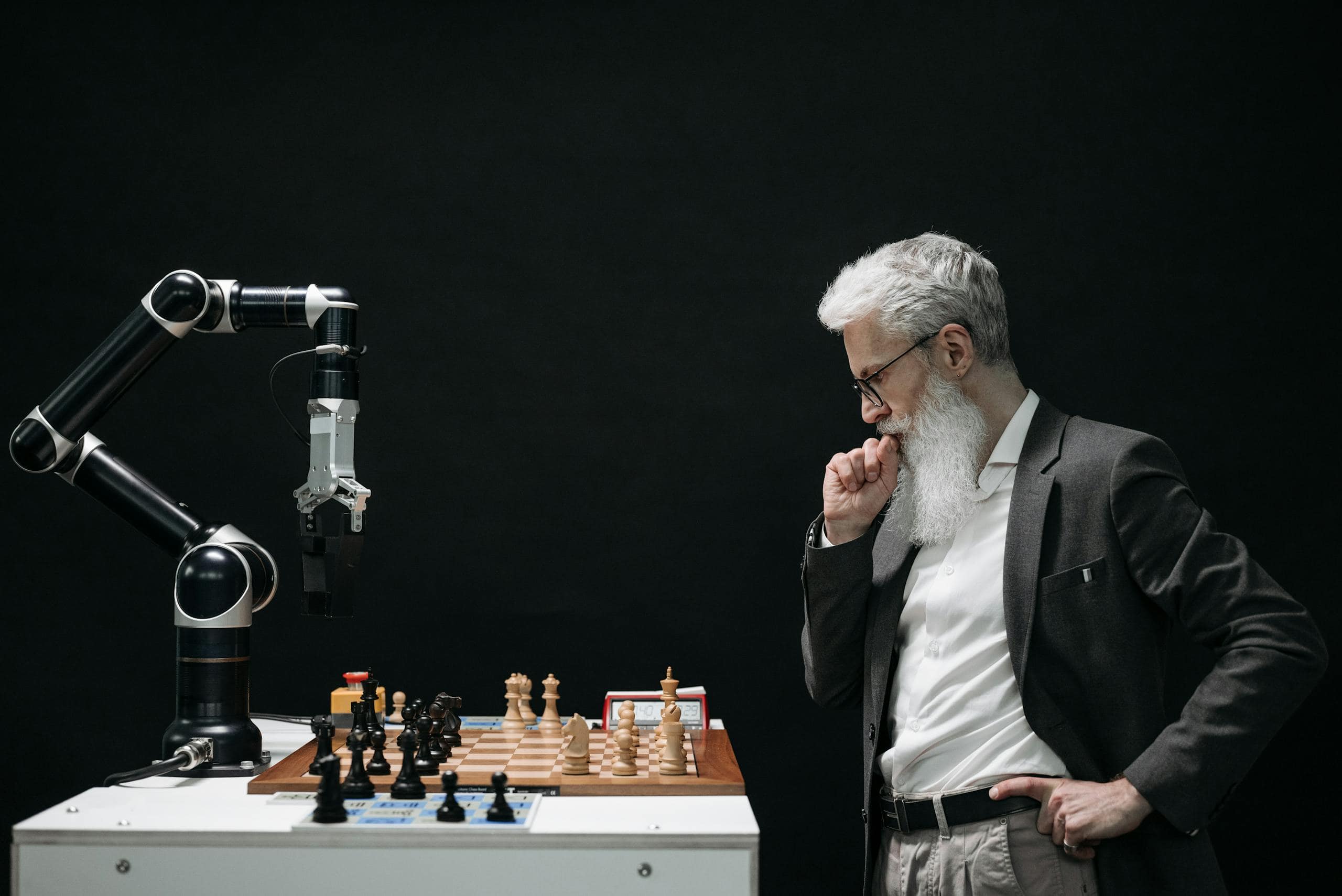 An elderly scientist contemplates a chess move against a robotic arm on a chessboard. US Government/Artificial Intelligence