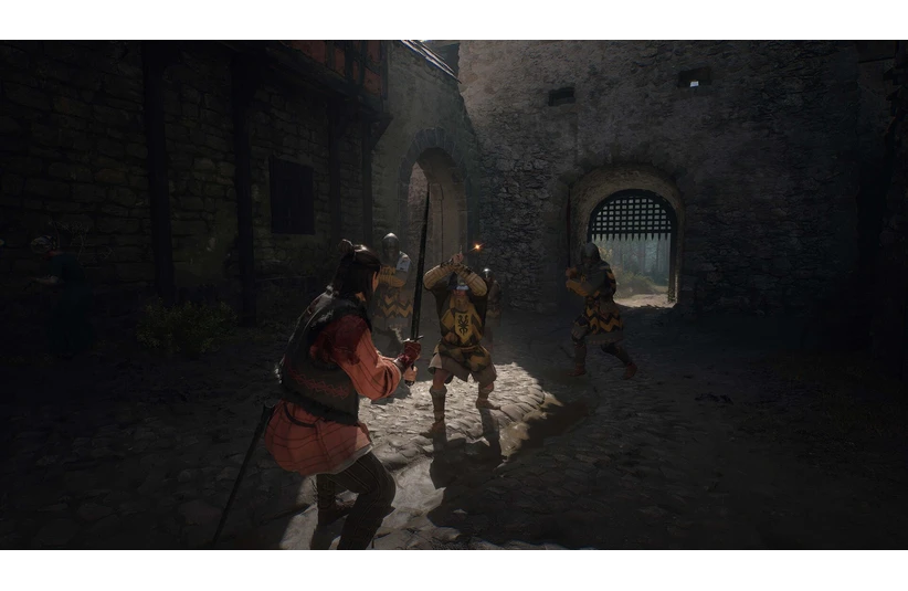 A medieval battle scene shows a warrior in red confronting two armored knights with swords in a shadowy stone courtyard, conveying tension and bravery. The Blood of Dawnwalker, Courtesy of Rebel Wolves and Bandai Namco Entertainment.