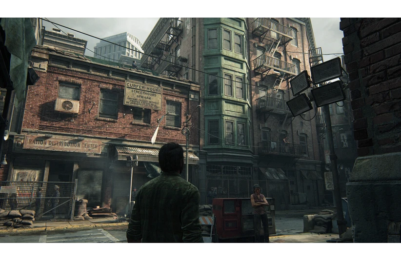 Joel, in a green jacket, stands in an abandoned, run-down city street. Surrounding are old brick buildings with fire escapes, creating a tense, post-apocalyptic atmosphere in The Last of Us
