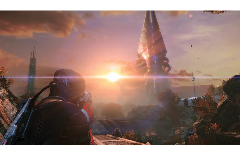 A futuristic soldier in Mass Effect in armor gazes at a towering alien structure. The setting sun casts a dramatic glow, highlighting a landscape of rocky terrain and buildings.